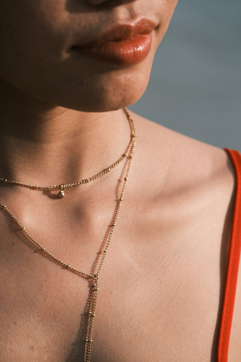 Close-up of a person wearing layered gold necklaces and a red top, with calm expression and soft lighting in a neutral background.