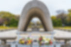Stone memorial arch in park with colorful flower tributes. Background shows trees, a distant dome, and gravel path. Calm atmosphere.
