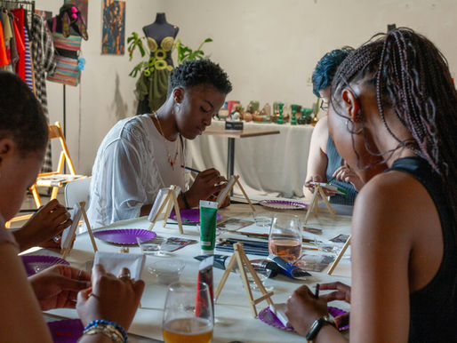 Young women paint at a craft table in an art studio, with clothes racks, mannequins, and supplies in the background.
