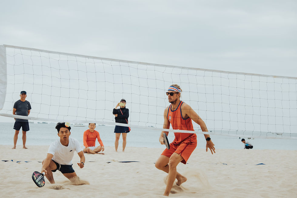 Two men play beach tennis intensely near a net; others watch. Overcast sky, sandy beach, and ocean in the background.