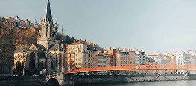 A riverside scene with a red bridge, historic buildings, and a large church with a tall spire, set against a hill under a clear sky.