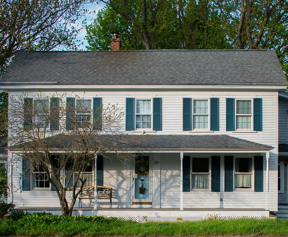 White two-story house with blue shutters, a front porch, and a tree with buds on it in the foreground.