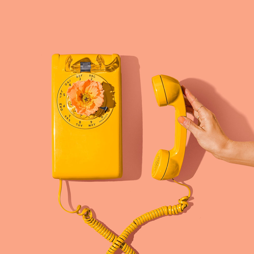 Yellow rotary phone with a floral dial on a pink background. Hand holds the yellow receiver, creating a playful and bright scene.
