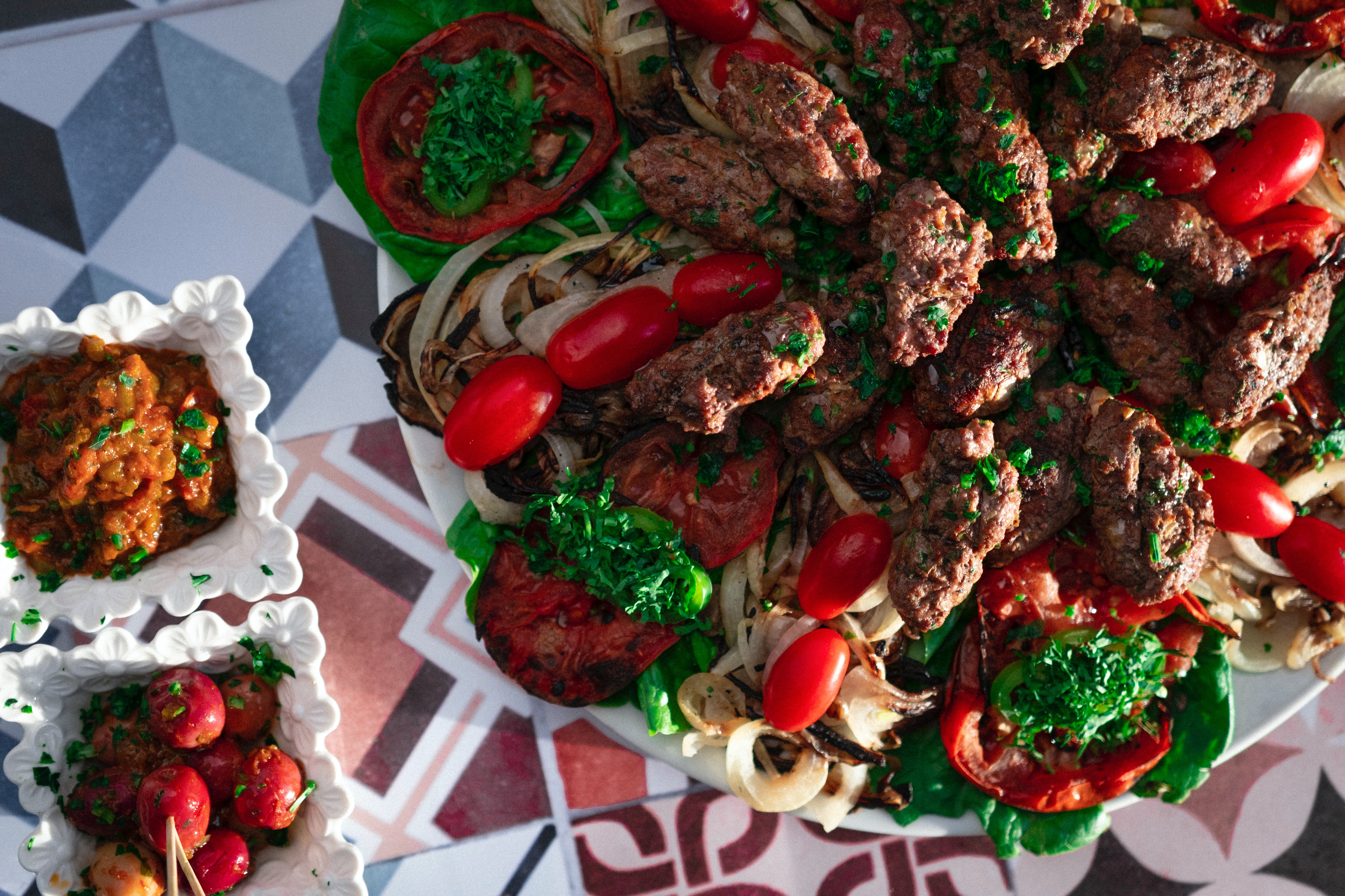 A colorful platter with grilled meat, cherry tomatoes, grilled onions, and tomato slices. Two bowls with side dishes on a patterned table representing Moroccan kefta dish.