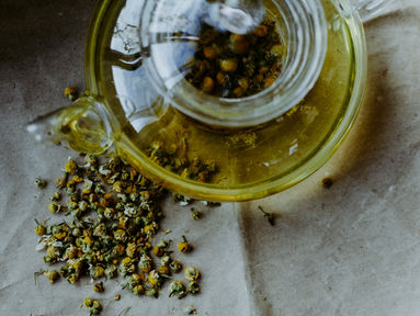 Glass teapot filled with chamomile tea next to scattered chamomile flowers on a textured surface. Earthy tones and serene mood.