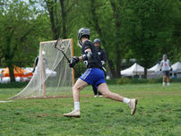 Lacrosse player in black jersey and blue shorts runs past the goal on a grassy field, with teammates and tents behind.