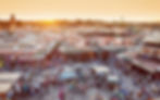 Marktplatz in Marrakesch bei Sonnenuntergang, voller Menschen, bunte Stände, orientalische Gebäude, geschäftige Atmosphäre, Gelb- und Rosatöne.