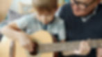 Child learning guitar with an older person. The child focuses on strumming strings, while the older person guides. Cozy indoor setting.