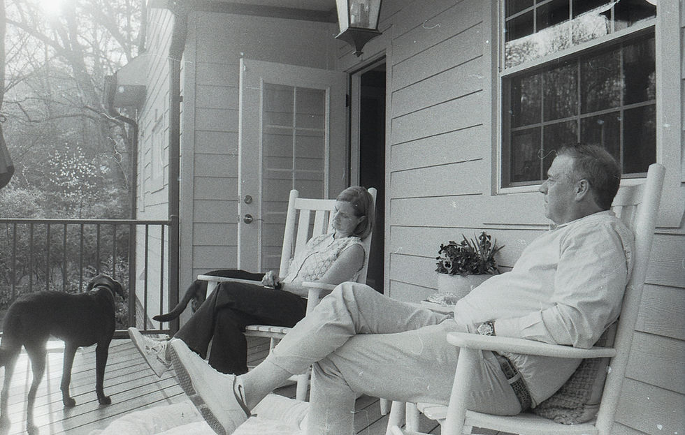 Two adults sit together in rocking chairs on a covered front porch with a dog nearby, creating a peaceful and welcoming scene that evokes feelings of comfort, connection, and home.