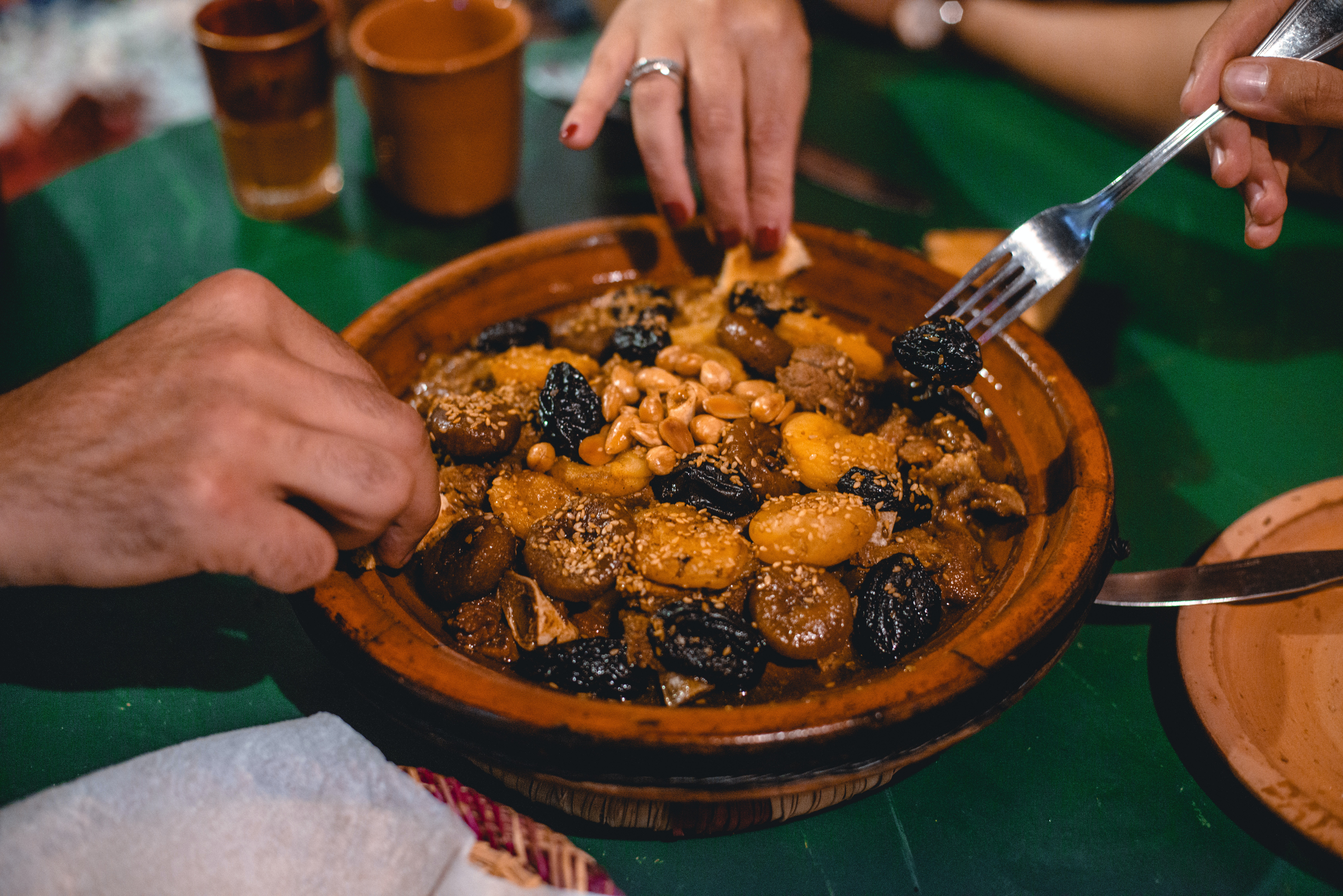 Moroccan tagine as a blog post cover for top foods in morocco. The picture showing a tagine full of meats and several hands surrounding it, taking a dip with bread pieces.
