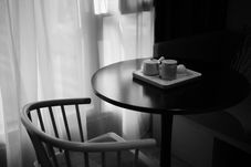 Monochrome café table by sheer curtains, with a chair and tea set on a tray, lit softly in a quiet, calm corner.