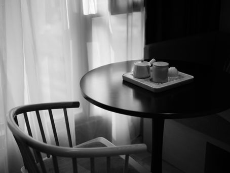Monochrome café table by sheer curtains, with a chair and tea set on a tray, lit softly in a quiet, calm corner.