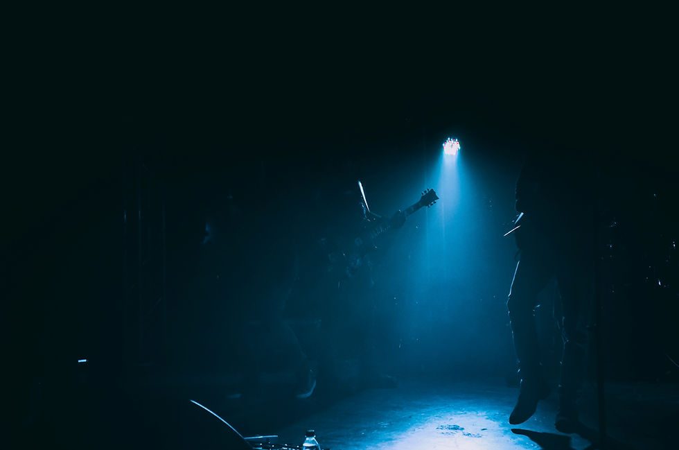 Image has dark background with a blue light shining on the leg of one person and part of a guitar held by a second person.