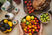 Top-down view of hands holding a bowl of colorful tomatoes, carrots, lemon and lime beside bread, avocados, and drink bottles.