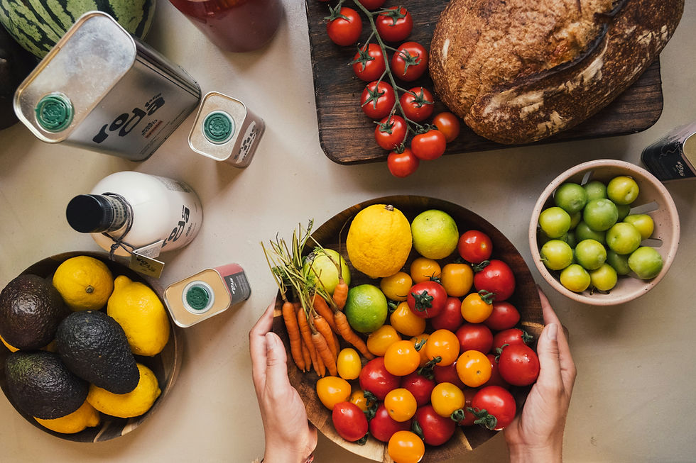 Top-down view of hands holding a bowl of colorful tomatoes, carrots, lemon and lime beside bread, avocados, and drink bottles.