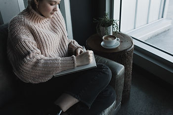 A woman wearing a pink sweater is sitting in a leather chair writing in a journal.