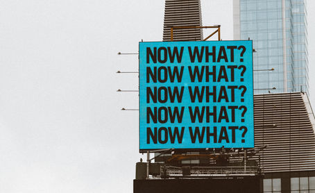 Blue billboard with black text "NOW WHAT?" repeated five times, set against a cityscape background with modern buildings. Overcast mood.
