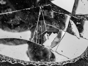 Broken mirror with intricate edge lies on the ground. Reflects fragmented images, creating a chaotic, disjointed pattern in black and white.