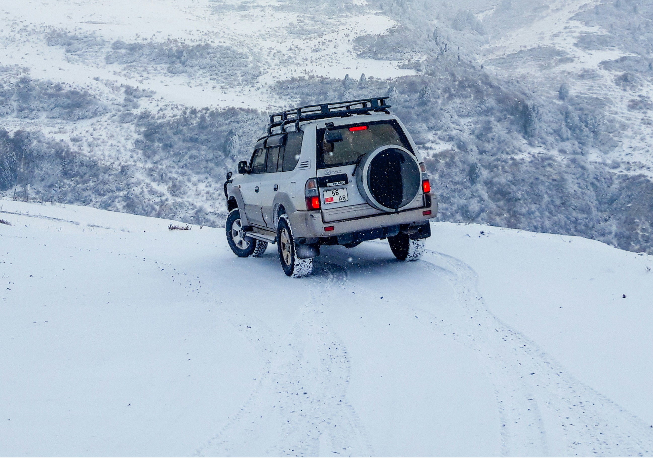 White SUV with roof rack on snowy mountain road in fog, tire tracks behind, red taillights and plate 56 AR visible.