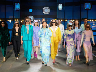 Fashion brand sales representative presenting SS27 fashion week spring summer 2027 collection to a retail buyer at a trade show booth, using a B2B fashion portal on a tablet device to display live inventory and confirm orders on the show floor during the SS27 season.