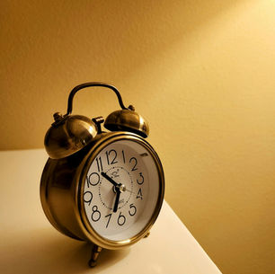 Brass twin-bell alarm clock on a white bedside table against a beige wall, lit by warm yellow light.
