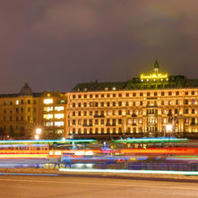 Illuminated grand hotel at night with light trails from passing boats. Buildings display warm lighting; sky is overcast. Text: Handelsbanken.