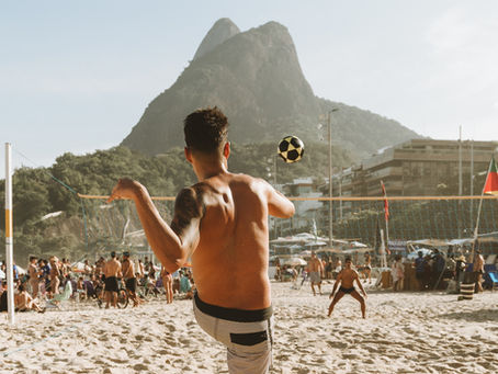 Shirtless man serves a ball on a sunny beach volleyball court, with crowds and a mountain backdrop.
