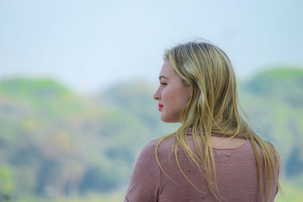 Jeune femme blonde de profil, en haut rose, regardant au loin devant un paysage flou de collines sous un ciel clair. Un Parcours PMA débute par un bilan de fertilité. Maman d'amour, accompagnement PMA et Fertilité en complément du parcours médical.