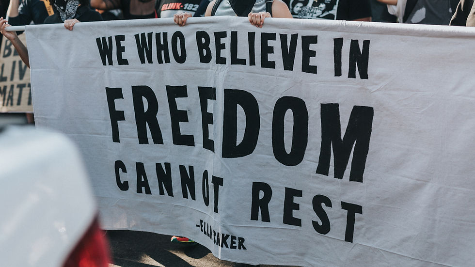 A group of people is holding a large white rectangular banner with bold black text that states, "We Who Believe in Freedom Cannot Rest". It is a famous quote by human rights activist Ella Baker.