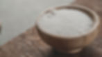 Wooden bowl filled with white sugar on a textured brown tabletop. Some sugar is spilled around, creating a rustic, cozy vibe.