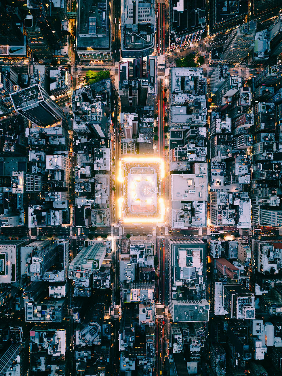 Vista aérea de uma cidade à noite, com prédios iluminados e tráfego nas ruas. Luzes brilhantes destacam uma área central quadrada.