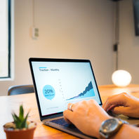 Hands type on a laptop showing Traction - Monthly and a 20% MoM Growth chart on a wooden desk with a small plant.