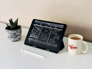 Tablet with wireframe design on screen, white stylus, mug with "I run on Caffeine" text, and potted plant on a white desk.