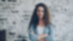 Woman with curly hair stands arms crossed before a white painted brick wall, wearing a blue cardigan and looking serious.