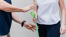 Trainer reaches toward a woman holding a green resistance band against a plain wall, suggesting a workout demo