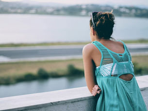 An over-the-shoulder view of a woman with her hair in a bun and sunglasses on her head, wearing a teal dress with a detailed macramé back. She leans against a stone balcony ledge in the Kanoni area of Corfu, looking out over the water toward the runway of Corfu International Airport. In the background, the iconic coastal runway spans parallel to the lagoon, surrounded by water and distant hazy green hills, capturing a classic vantage point used by tourists for plane spotting.