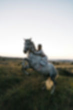 Woman in a floral dress hugs a rearing white horse in a grassy field at sunset, with rolling hills in the background.