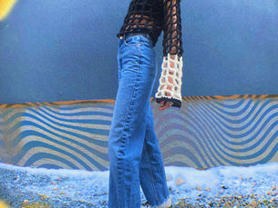Gen z female with long, wavy dark brown hair is posing stylishly in front of a wall painted with blue and yellow graffiti. She is wearing a black and white crochet top with long blue jeans.