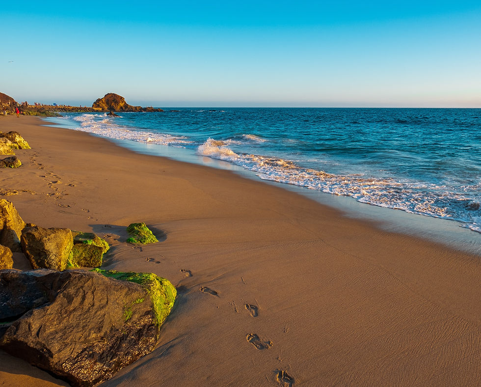 A serene beach at sunset with gentle waves, footprints in the sand, and rocks with green moss. The horizon is clear and blue.