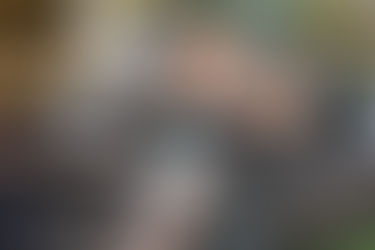 Man in a straw hat carving wood with a knife near a campfire. Warm flames and earthy tones create a rustic outdoor setting.