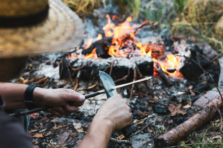 Man in a straw hat carving wood with a knife near a campfire. Warm flames and earthy tones create a rustic outdoor setting.