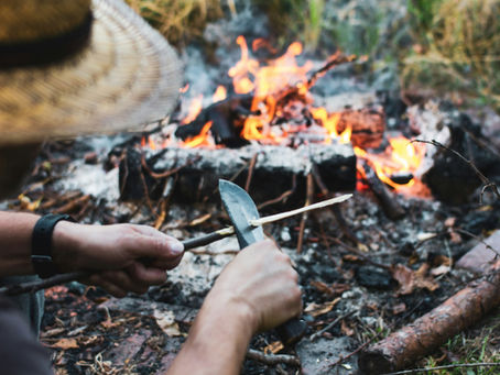 Man in a straw hat carving wood with a knife near a campfire. Warm flames and earthy tones create a rustic outdoor setting.