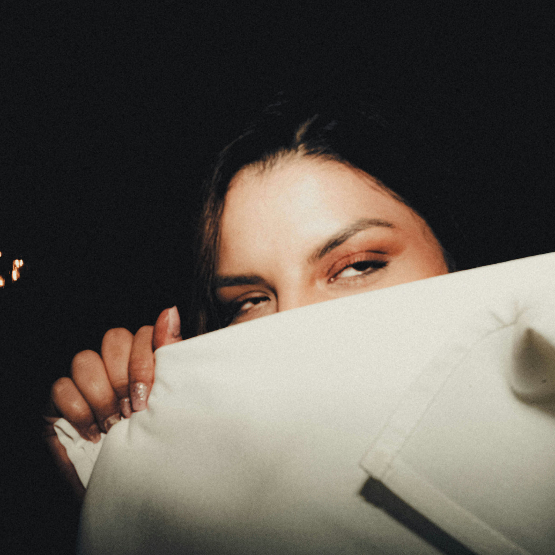 Close-up of a woman peeking over a white napkin in a dark room, eyes half-closed, with warm lights and a sleepy mood