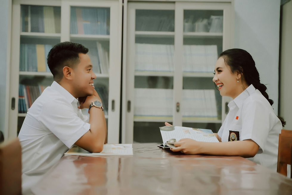 Two students in white uniforms laugh across a table in a classroom, reading books with shelves behind them.