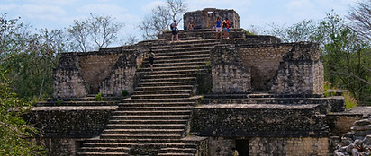 Visitors stand atop the ancient stone steps of a Mayan pyramid structure surrounded by trees under a partly cloudy sky.