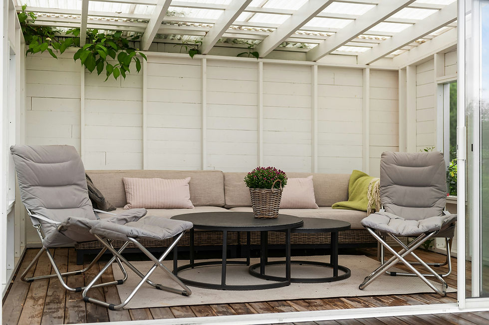 Cozy patio with beige sofa, pink cushions, two gray chairs, and a basket of flowers on a black table. Wooden floor, white walls, leafy decor.