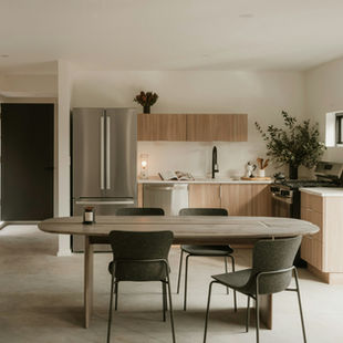 Minimalist kitchen with wooden cabinets, gray table, and chairs. Open book, plant, candle, and stainless steel fridge in bright, airy room.