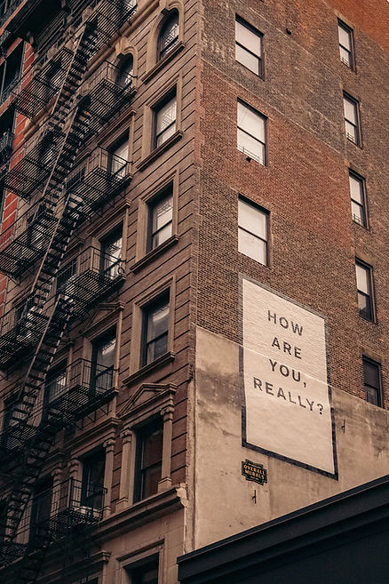 Brick building with fire escapes and a large mural reading "HOW ARE YOU, REALLY?" in black text on a white background. Urban setting.