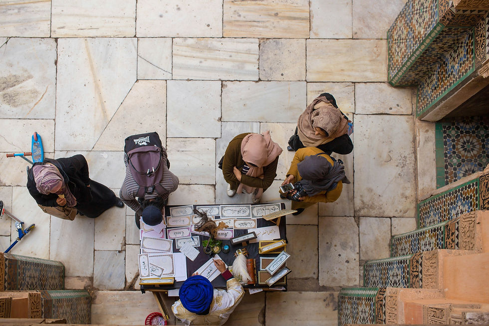 People gather around a table with calligraphy on paper, in a tiled courtyard. Notable colors: earth tones, blue, and green.