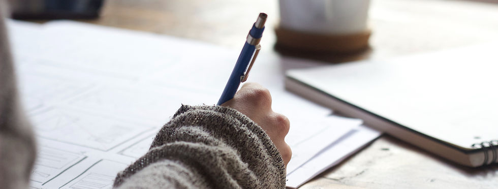 A person in a gray sweater writes on paper with a blue pen at a wooden table.